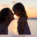 A woman and young child touch foreheads beside the water at sunset.