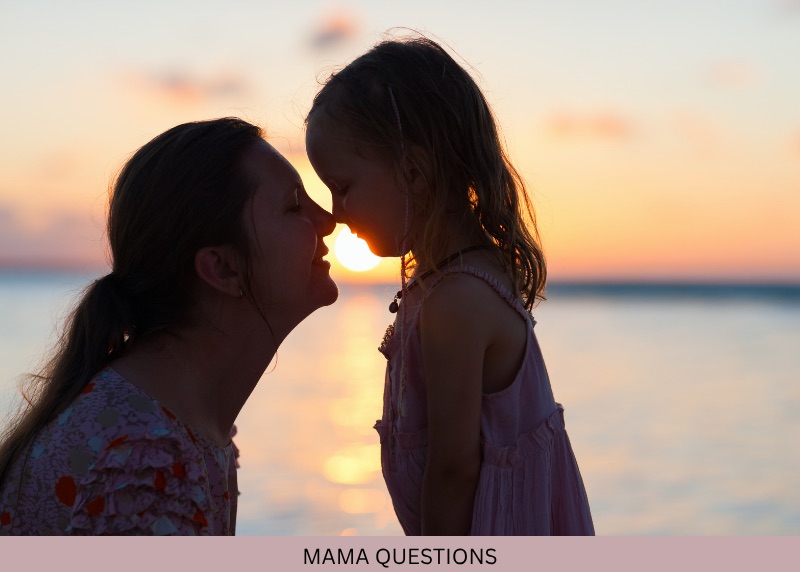 A woman and young child touch foreheads beside the water at sunset.